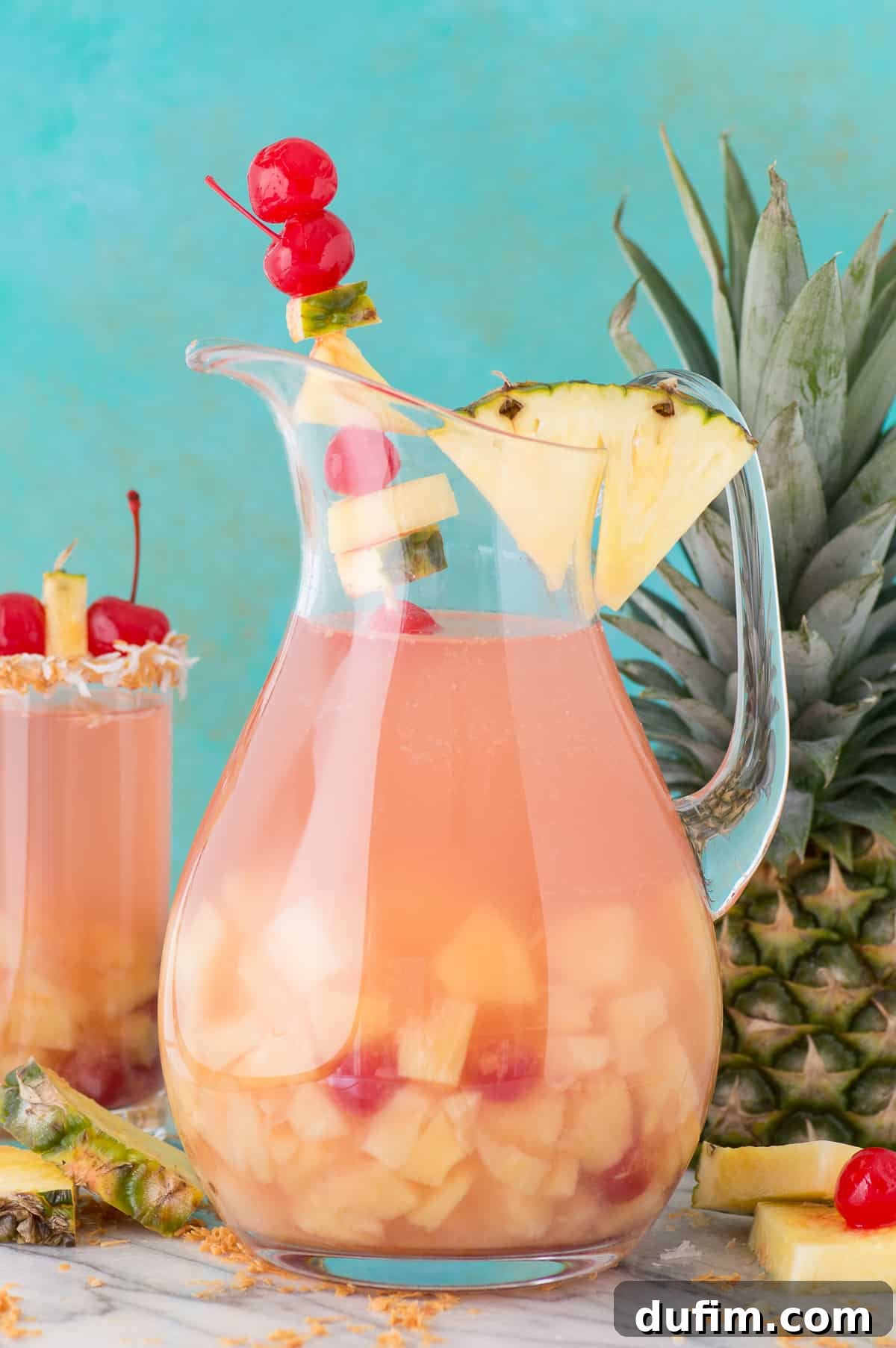 A large glass pitcher filled with colorful Pina Colada Sangria, brimming with pineapple chunks and maraschino cherries.