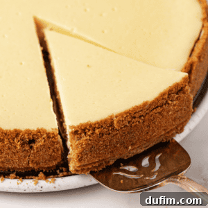 A close-up of a classic cheesecake slice with a graham cracker crust on a plate, next to the whole cheesecake, served with a metal cake server.