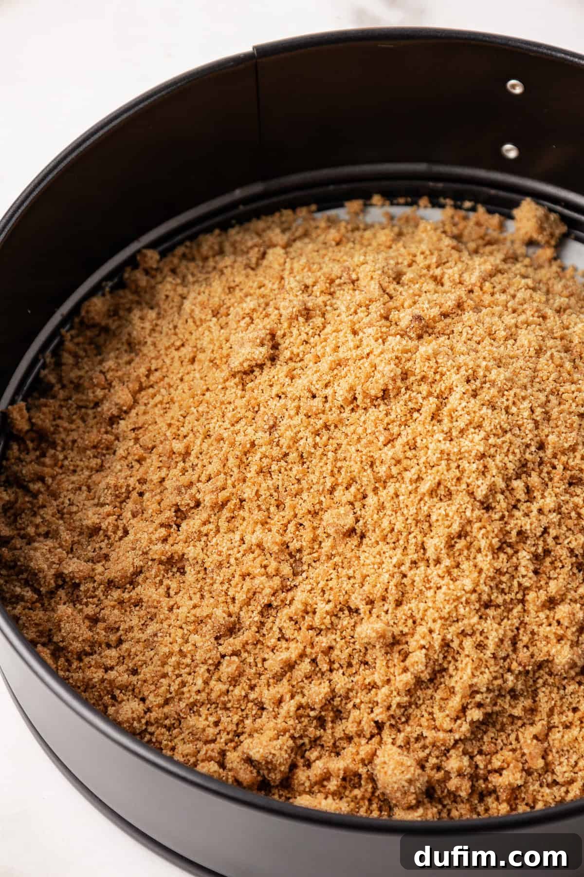 The graham cracker crust mixture being pressed firmly into the bottom of a springform pan using the base of a glass.
