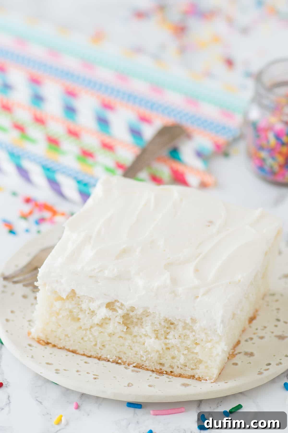 A perfectly baked slice of white cake on a white plate, complemented by colorful drinking straws in the background, ready to be enjoyed.