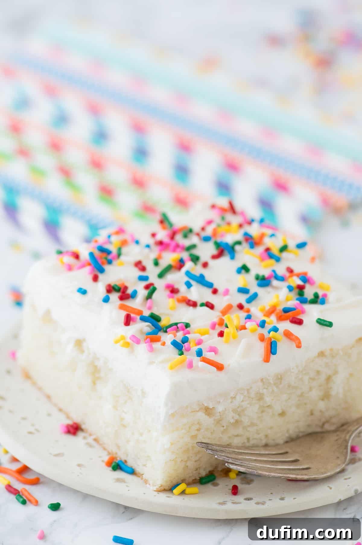 A slice of our easy white cake with a generous layer of white frosting and festive rainbow sprinkles, presented on a simple white plate.