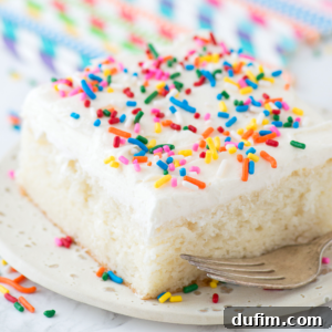 A slice of easy white cake from box with white frosting and colorful rainbow sprinkles sits on a white plate, with a fork beside it.