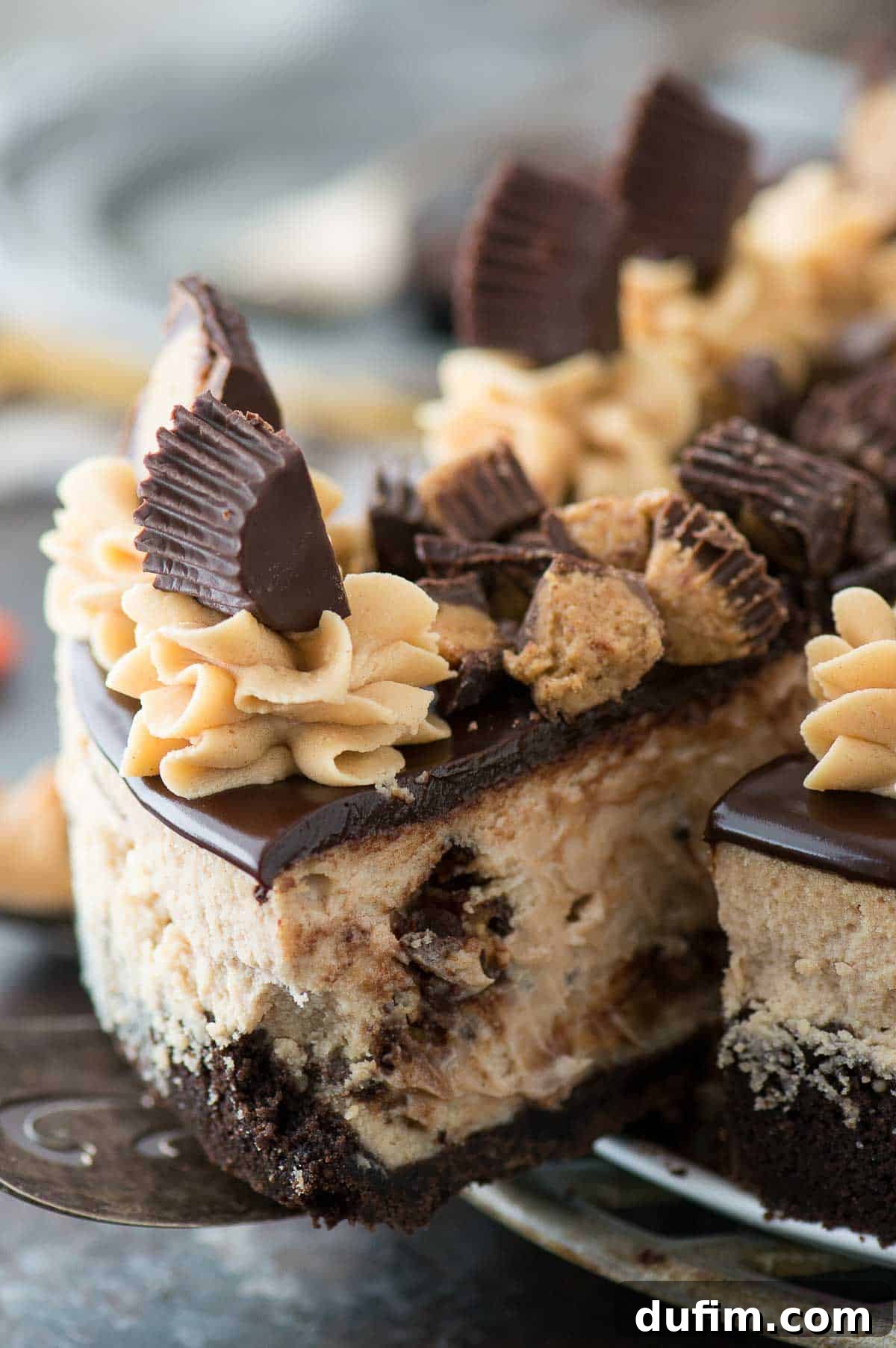 A close-up shot of a magnificent slice of Reese's cheesecake, featuring its layers of chocolate crust, peanut butter filling with candy pieces, and a generous topping of chocolate ganache and more Reese's cups on a metal plate.