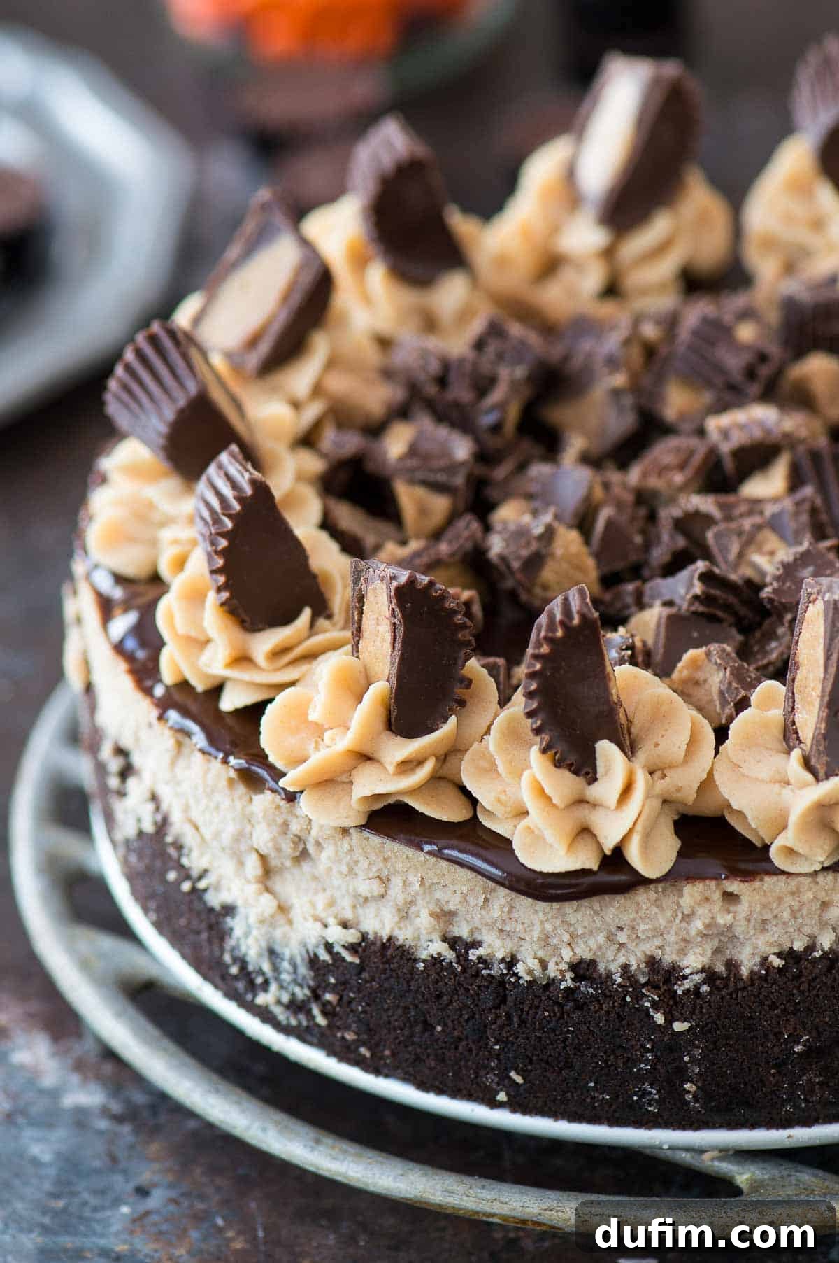 A stunning Reese's cheesecake, beautifully garnished with chocolate ganache and an abundance of Reese's cups, presented on a shiny metal plate.