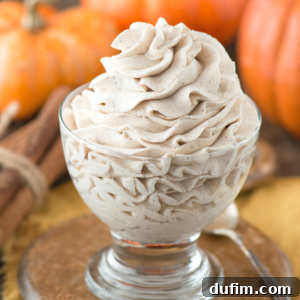 A beautifully presented glass dessert cup filled with swirled pumpkin whipped cream, garnished with elegant cinnamon sticks, with autumn pumpkins softly blurred in the background.