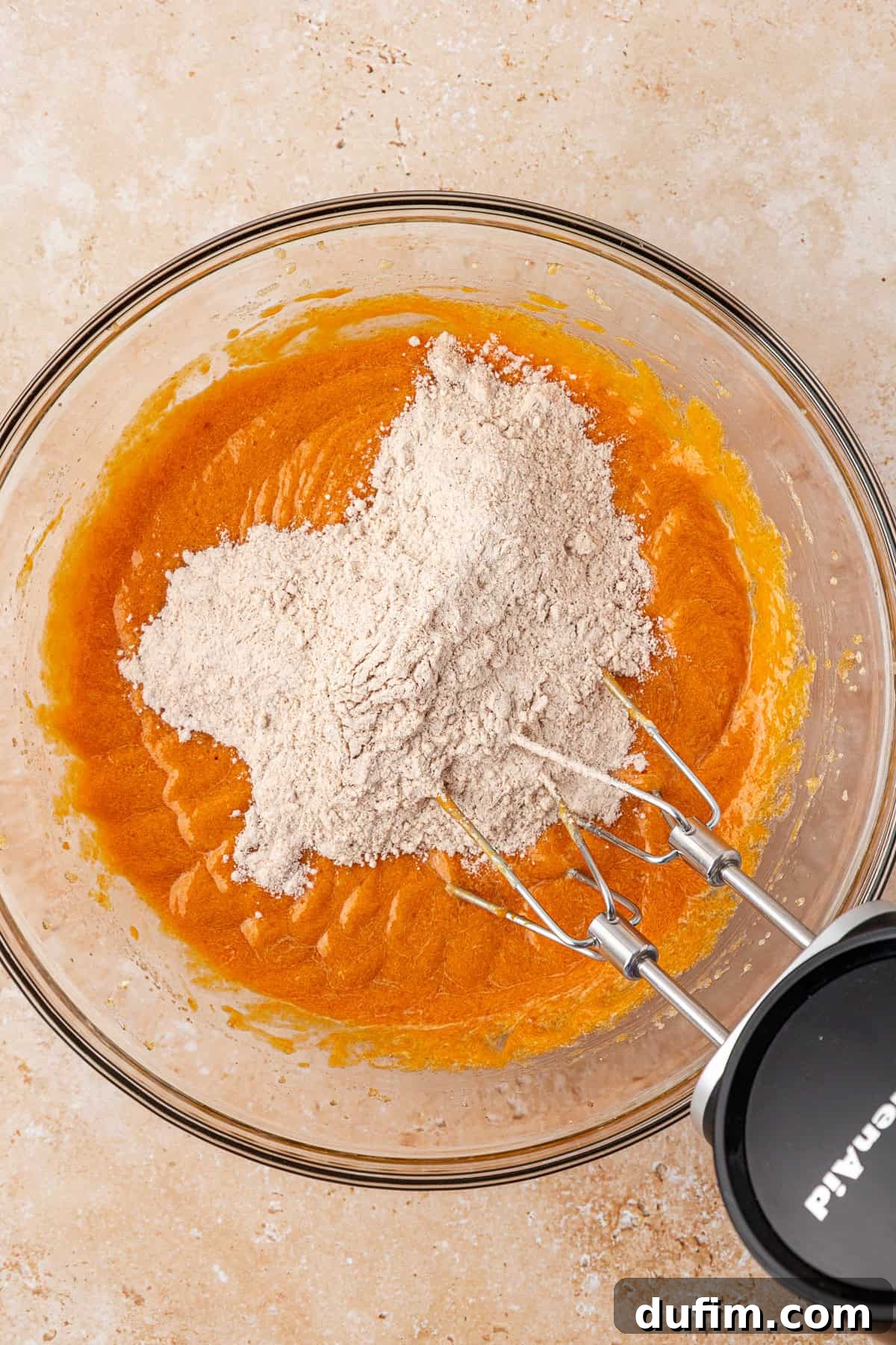 A glass bowl with orange batter and a mound of flour on top, partially mixed with an electric hand mixer resting inside the bowl, on a light-colored countertop.