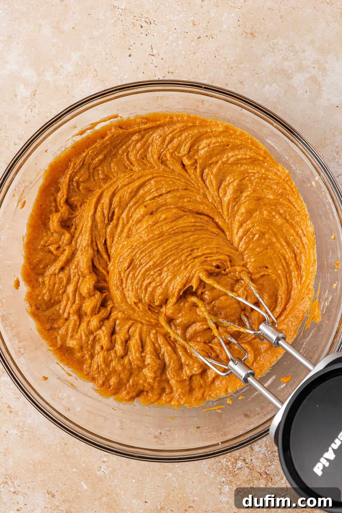 A glass bowl filled with pumpkin-colored batter being mixed with an electric hand mixer on a light brown countertop.