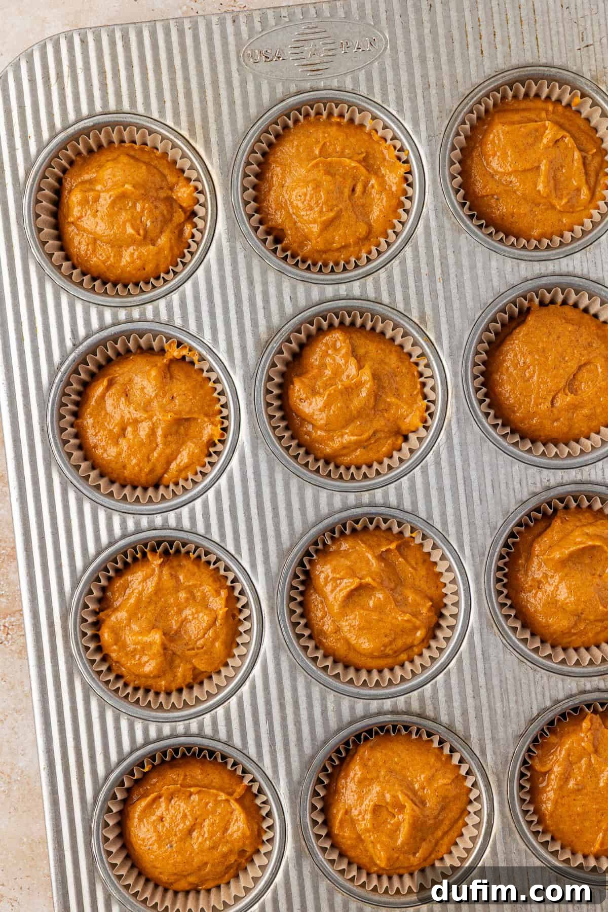 A metal muffin tray filled with twelve paper liners, each containing unbaked orange muffin batter. The tray is on a light-colored surface.