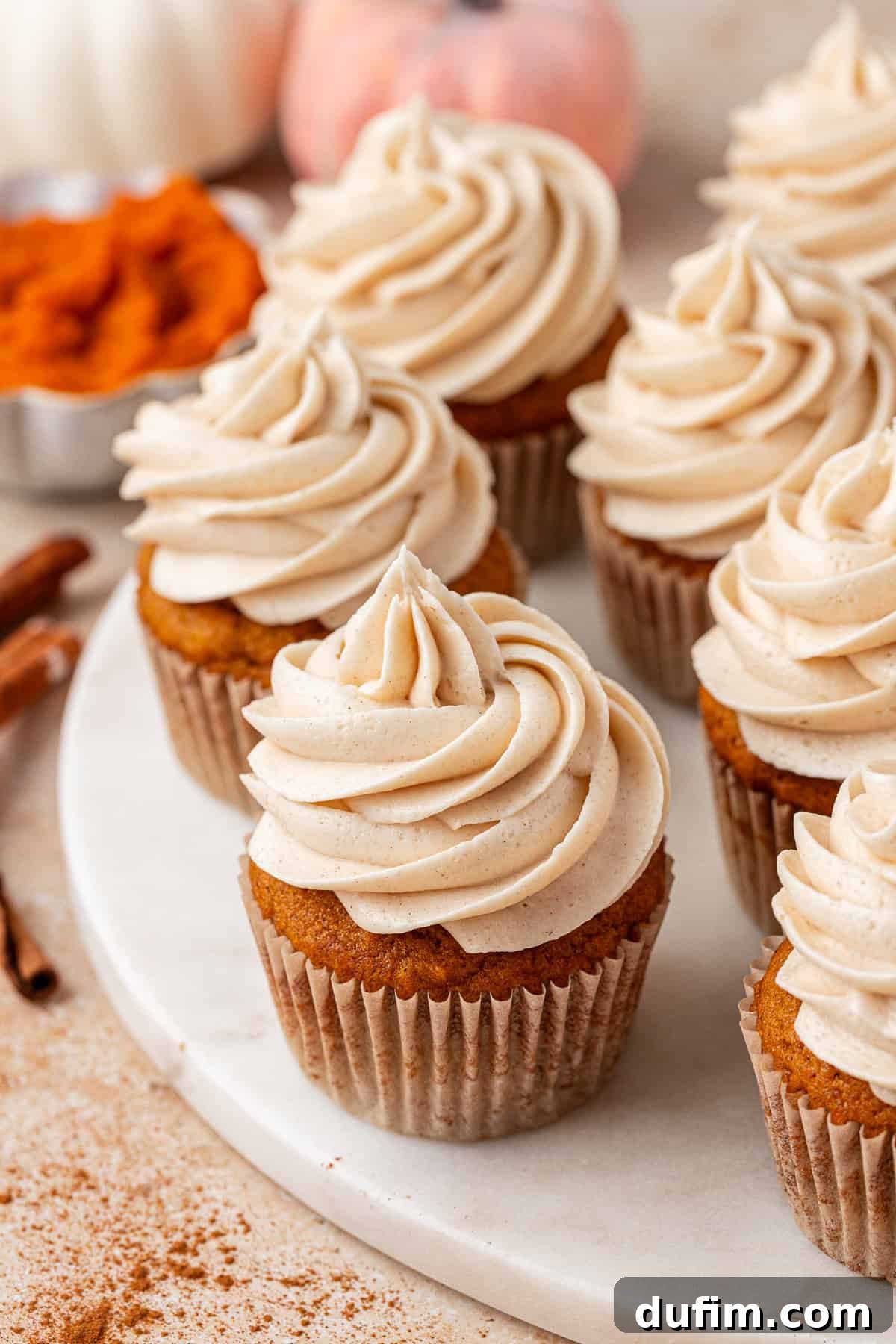 Pumpkin cupcakes with swirled cinnamon frosting sit on a white marble surface, surrounded by cinnamon sticks and a bowl of pumpkin puree in the background.