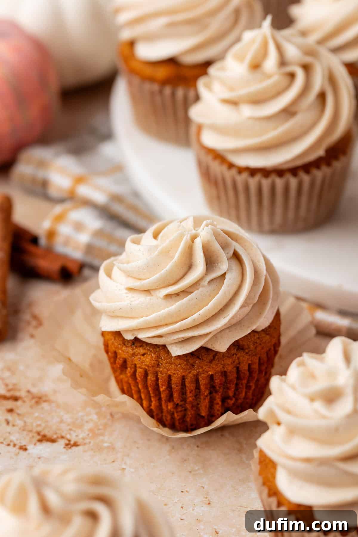 A close-up of pumpkin cupcakes topped with swirls of creamy frosting, set on a light surface with fall-themed decor and cinnamon sticks in the background.