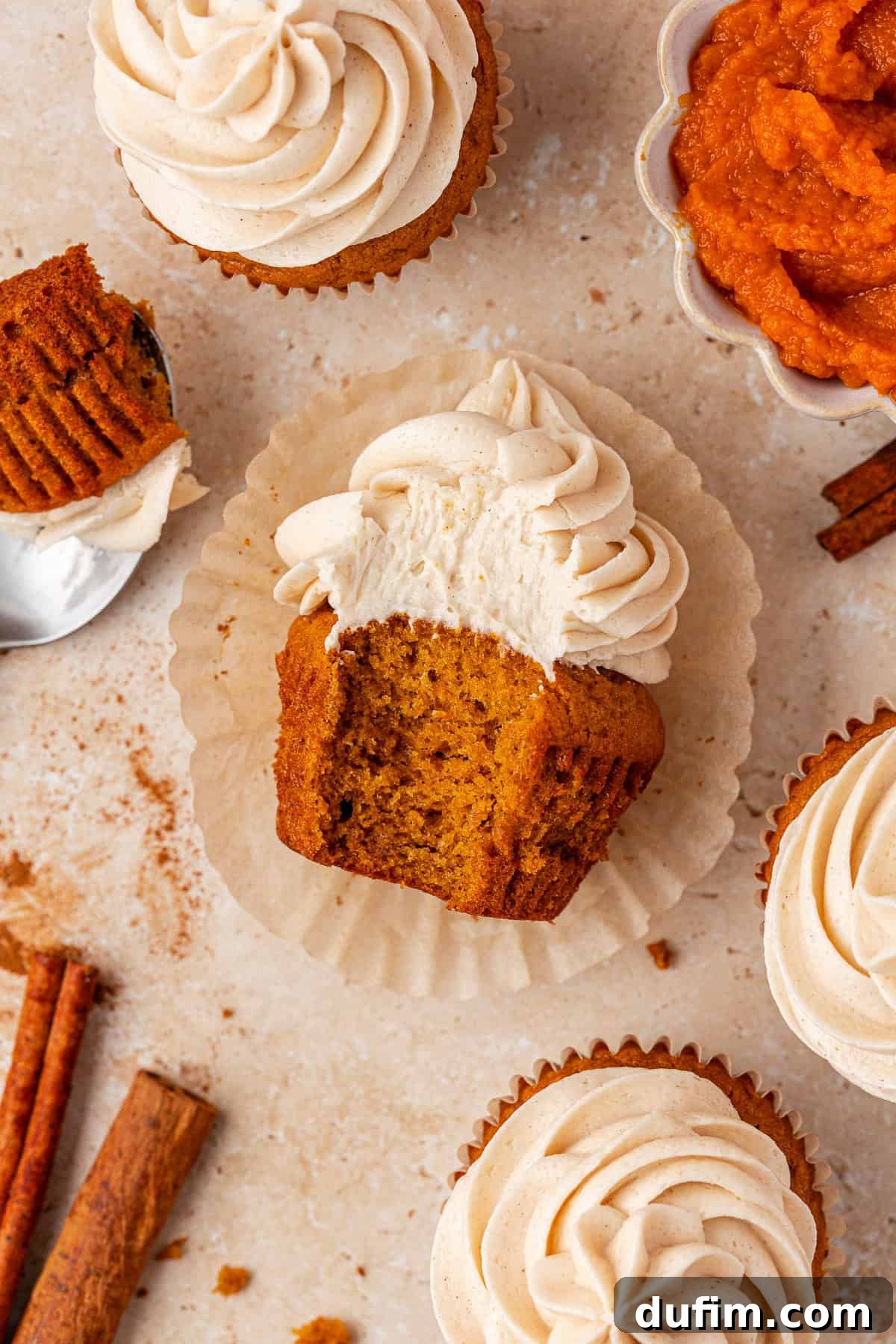 A pumpkin cupcake with swirled frosting sits on a wrapper, showing a bite taken out. Surrounding it are whole cupcakes, a bowl of pumpkin puree, cinnamon sticks, and scattered crumbs on a light surface.