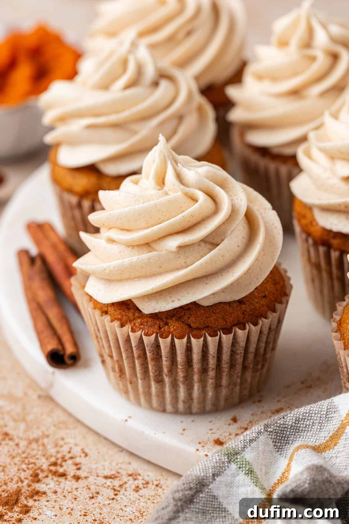 A close-up of pumpkin cupcakes with swirls of creamy frosting on top, arranged on a white plate. Two cinnamon sticks and a sprinkle of cinnamon powder are nearby. A checkered napkin and a bowl of orange filling are in the background.
