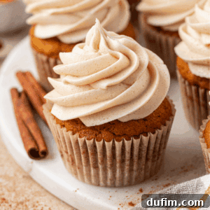 A pumpkin cupcake with swirled cinnamon frosting sits on a white plate, with cinnamon sticks nearby and other cupcakes in the background.