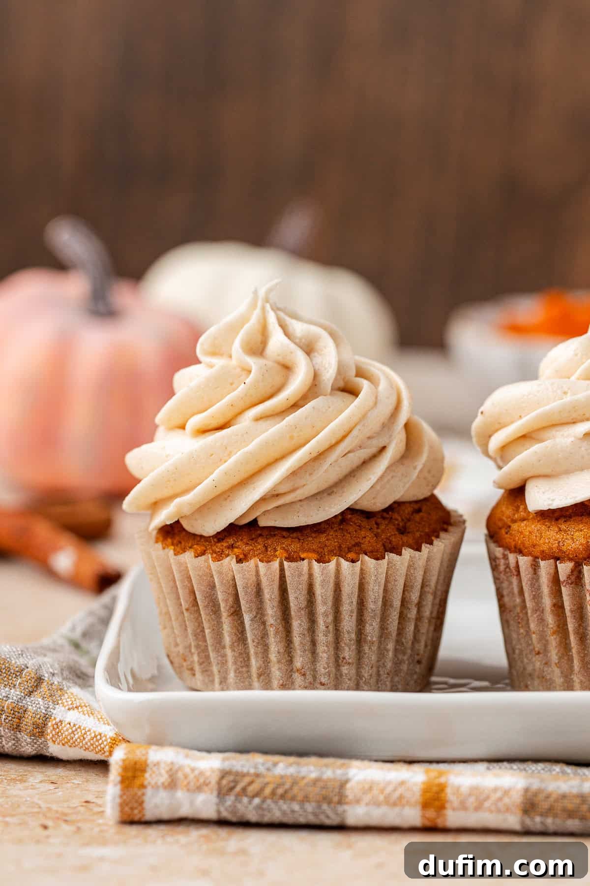 A pumpkin cupcake topped with swirled cream frosting sits on a white plate, with another cupcake beside it. A plaid napkin, cinnamon sticks, and blurred pumpkins are in the background.