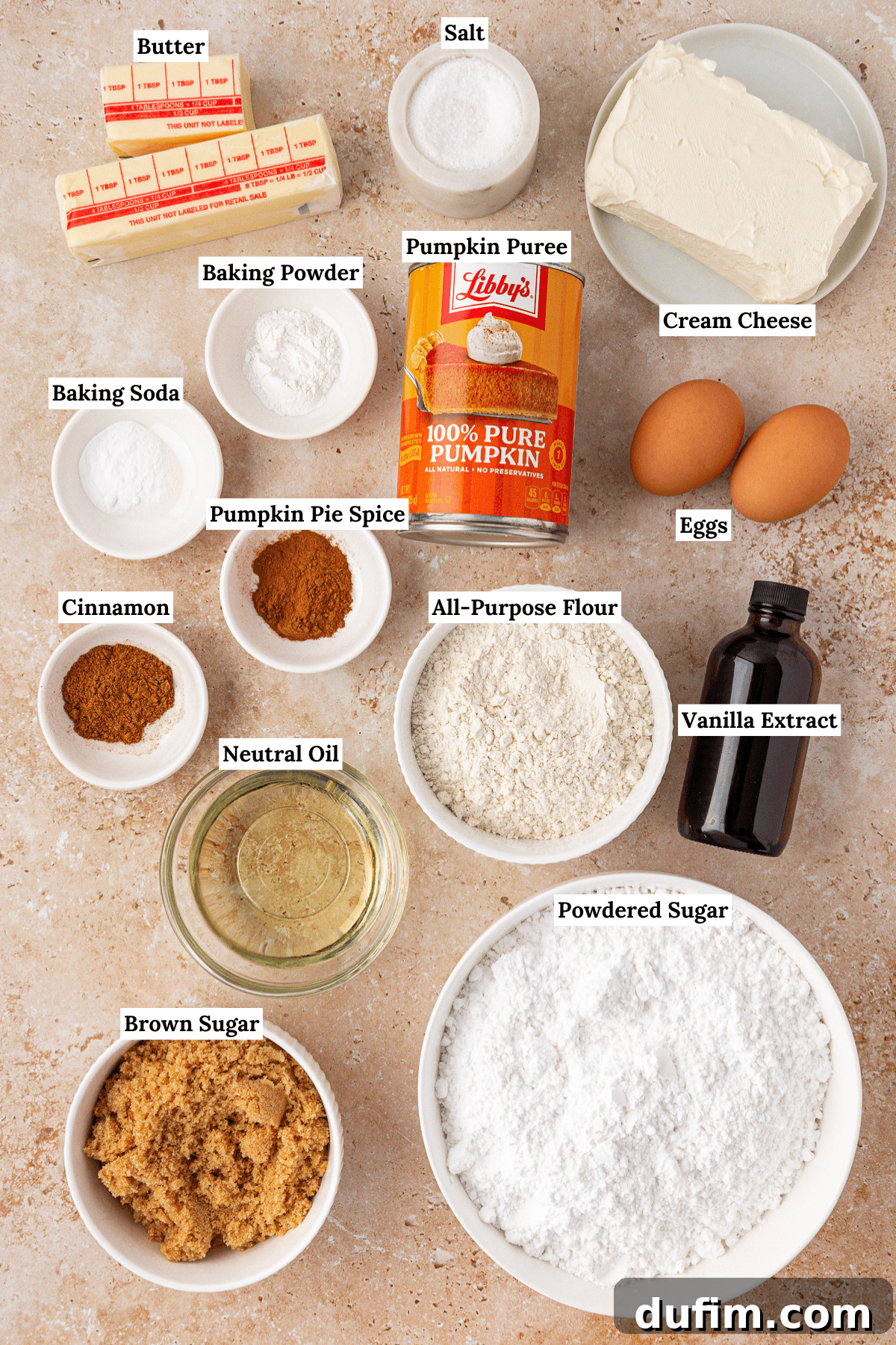Labeled baking ingredients on a countertop, including butter, salt, cream cheese, eggs, vanilla extract, powdered sugar, brown sugar, neutral oil, all-purpose flour, pumpkin puree, pumpkin pie spice, cinnamon, baking soda, and baking powder.