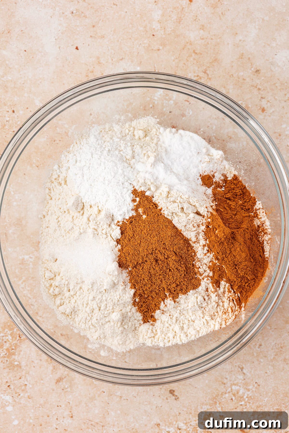 A glass bowl filled with unmixed dry baking ingredients, including flour, sugar, baking powder, and ground spices, on a beige countertop.