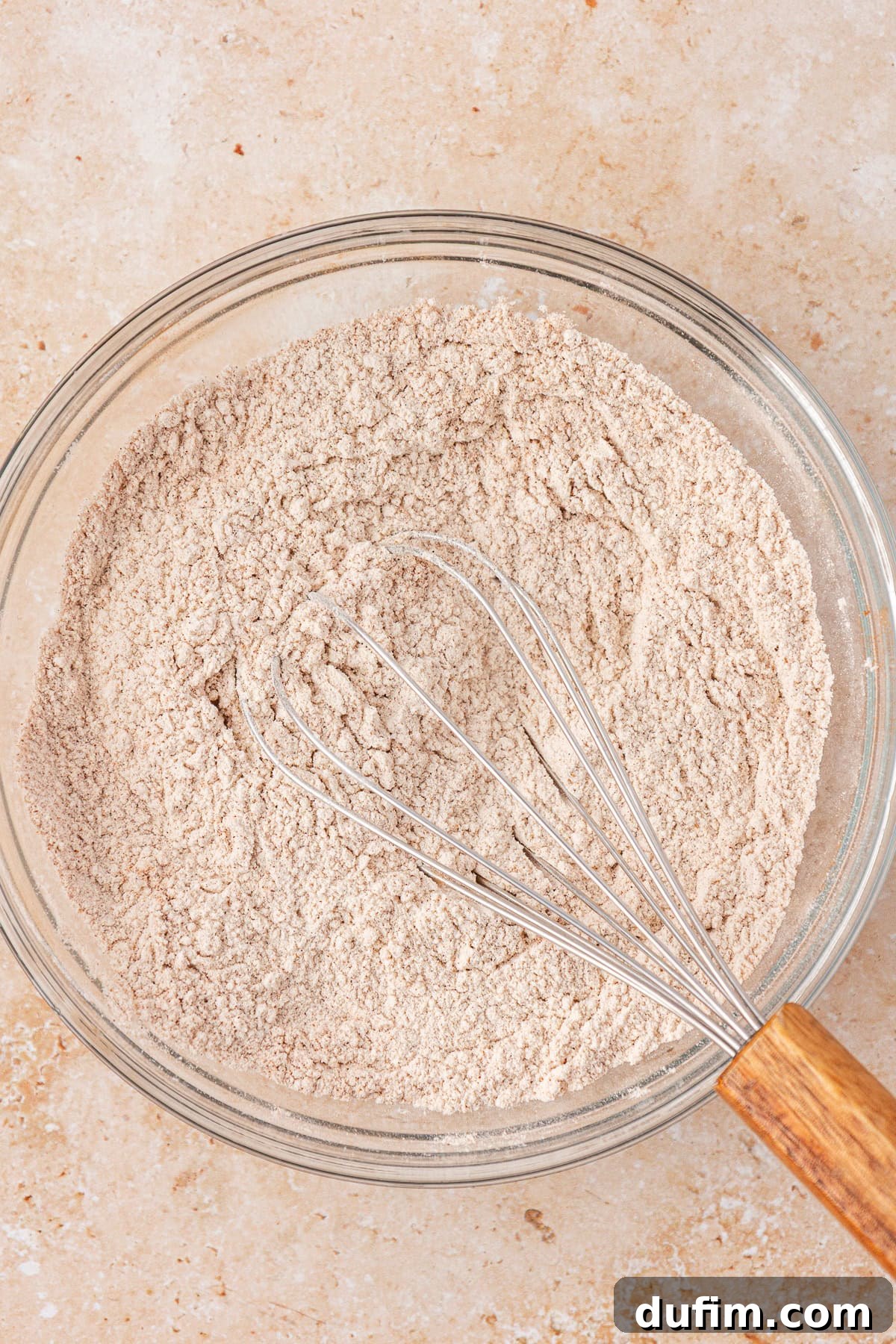 A glass bowl filled with a light brown flour mixture and a metal whisk with a wooden handle, resting on a light beige countertop.