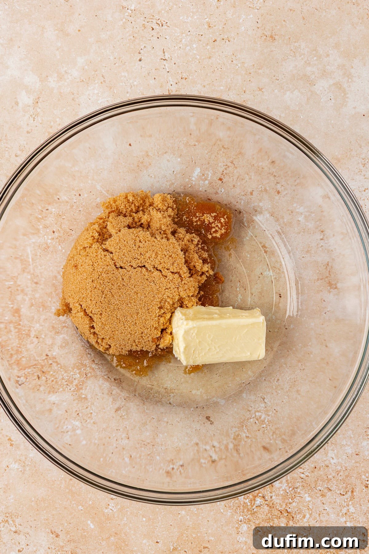 A glass mixing bowl containing a mound of brown sugar and a chunk of butter on a light beige countertop.