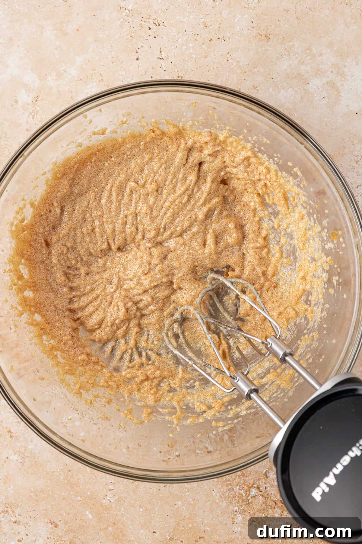 A glass mixing bowl with creamed brown sugar and butter, partially mixed with an electric hand mixer resting inside the bowl on a beige countertop.