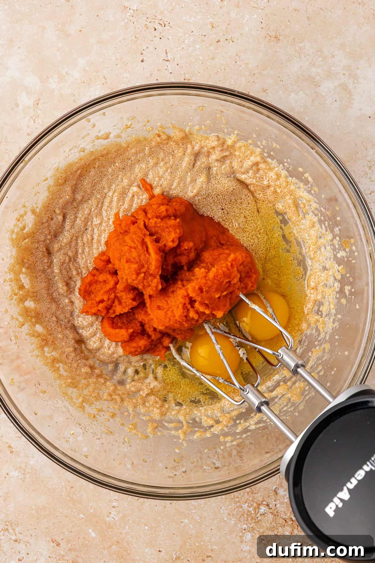 A glass bowl with mixed brown sugar and butter, three raw eggs, and a mound of pumpkin puree, with a hand mixer positioned inside, on a beige countertop.