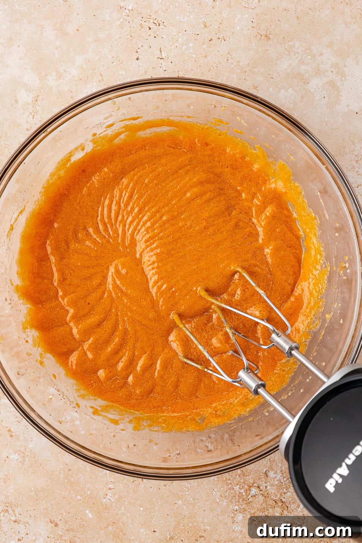 A glass mixing bowl filled with orange batter sits on a light countertop. An electric hand mixer with two beaters rests inside the bowl, showing swirled patterns in the smooth batter.