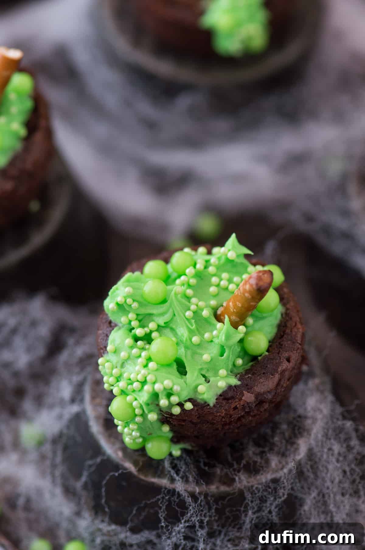 Mini brownie bites filled with vibrant green frosting and sparkling green sprinkles, artfully arranged to resemble bubbling witch's cauldrons, set against a spooky Halloween backdrop.