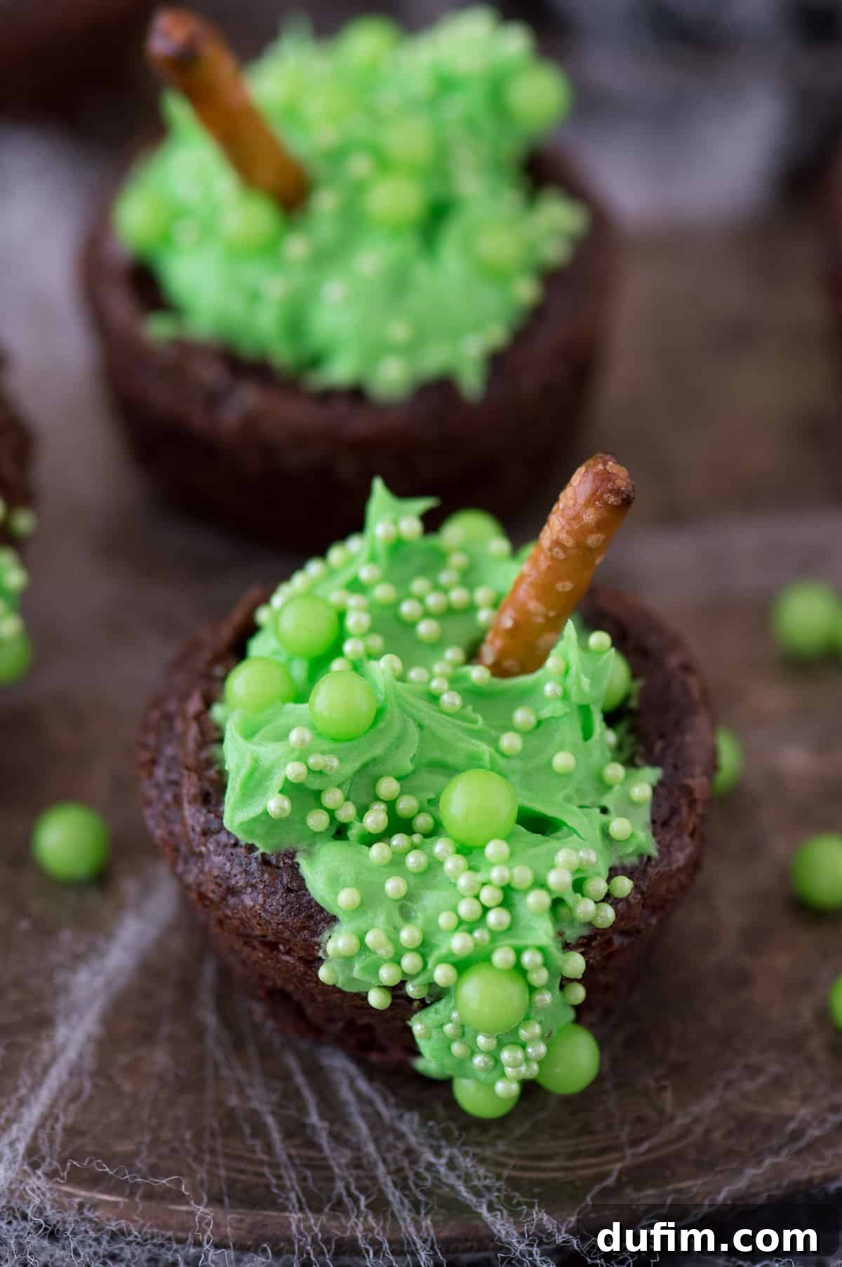 Close-up of mini brownie bites styled as witch's cauldrons, featuring gooey green frosting and a mix of large and small green sprinkles, with a small pretzel stick acting as a stirring spoon.