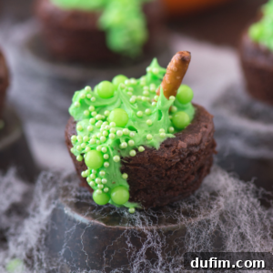 A Halloween brownie bite decorated to look like a bubbling witch's cauldron, with green frosting, green candy sprinkles, and a pretzel stick as a stirring spoon, surrounded by fake cobwebs.