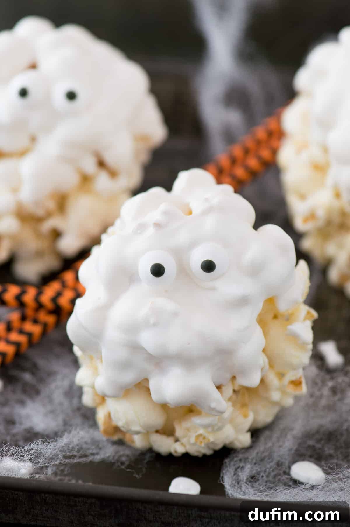 Three completed ghost popcorn balls with candy eyeballs resting on a silicone baking mat, ready to be served or stored. The background shows more Halloween decorations.