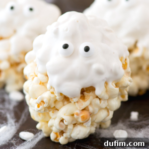 A ghost popcorn ball is coated in white frosting and decorated with two candy eyes, resembling a spooky Halloween popcorn ball. The treat sits on a black surface with wispy white strands that look like spider webs.