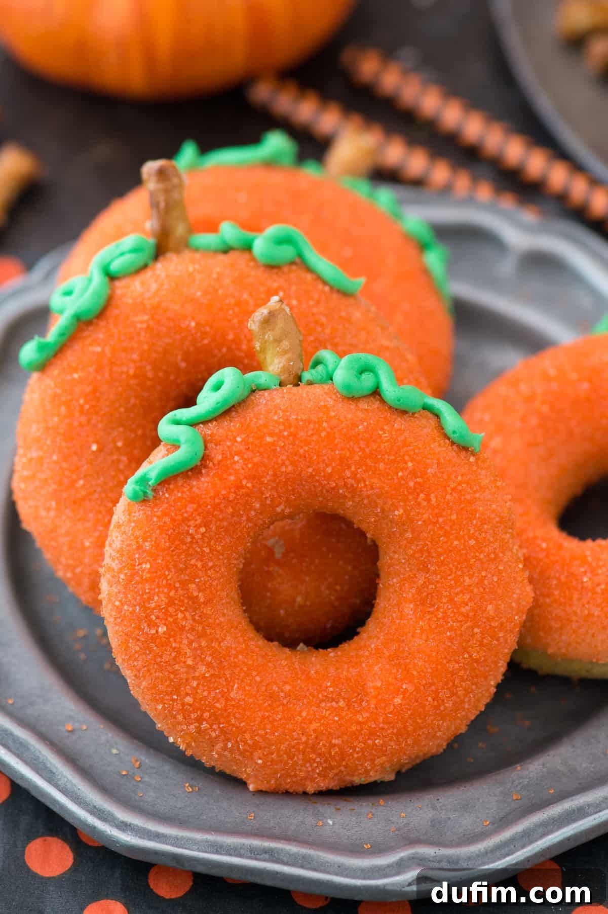 Golden Pumpkin Puffs 2 Two beautifully decorated pumpkin-shaped donuts on a cooling rack, garnished with orange sanding sugar, green frosting 'vines,' and pretzel 'stems'.