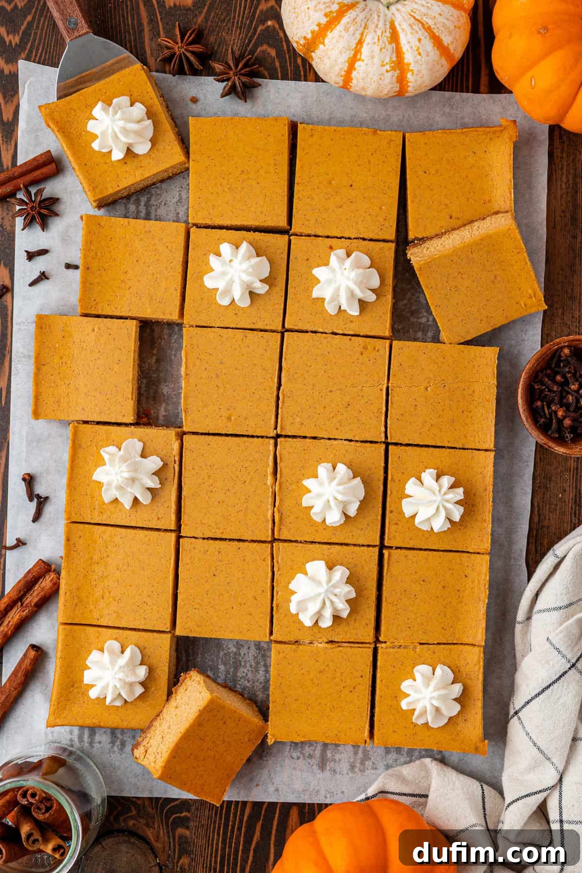 Spiced Pumpkin Cheesecake Squares 2 Top-down view of pumpkin bars cut into squares on parchment paper, some topped with whipped cream, surrounded by whole mini pumpkins, cinnamon sticks, star anise, and cloves on a wooden table. These easy-to-make pumpkin cheesecake bars are baked in a 9x13 pan with a simple graham cracker crust and a luscious 7-ingredient filling, making them perfect for any autumn celebration.