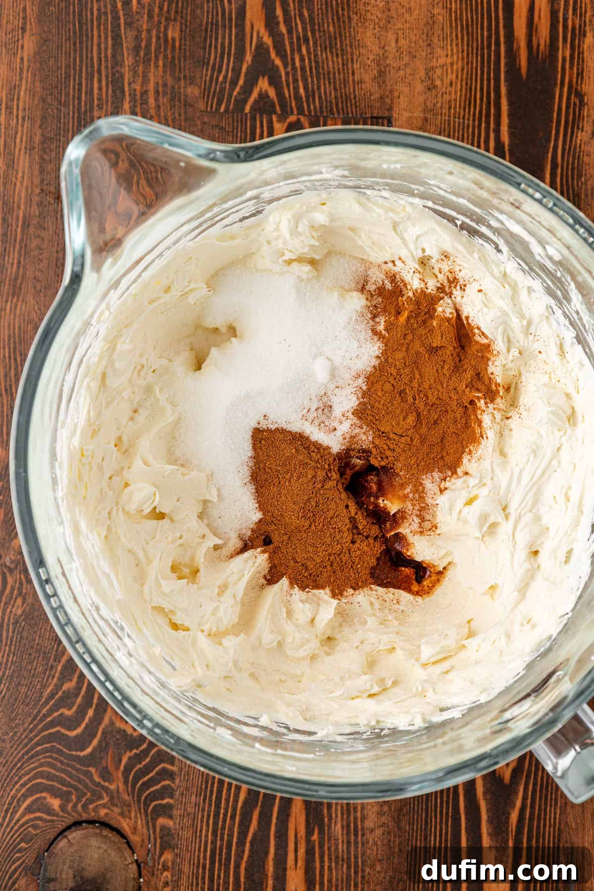 Spiced Pumpkin Cheesecake Squares 12 A glass mixing bowl on a wooden surface contains whipped cream cheese topped with granulated sugar, ground cinnamon, and vanilla extract, ready to be mixed together for the pumpkin cheesecake filling.