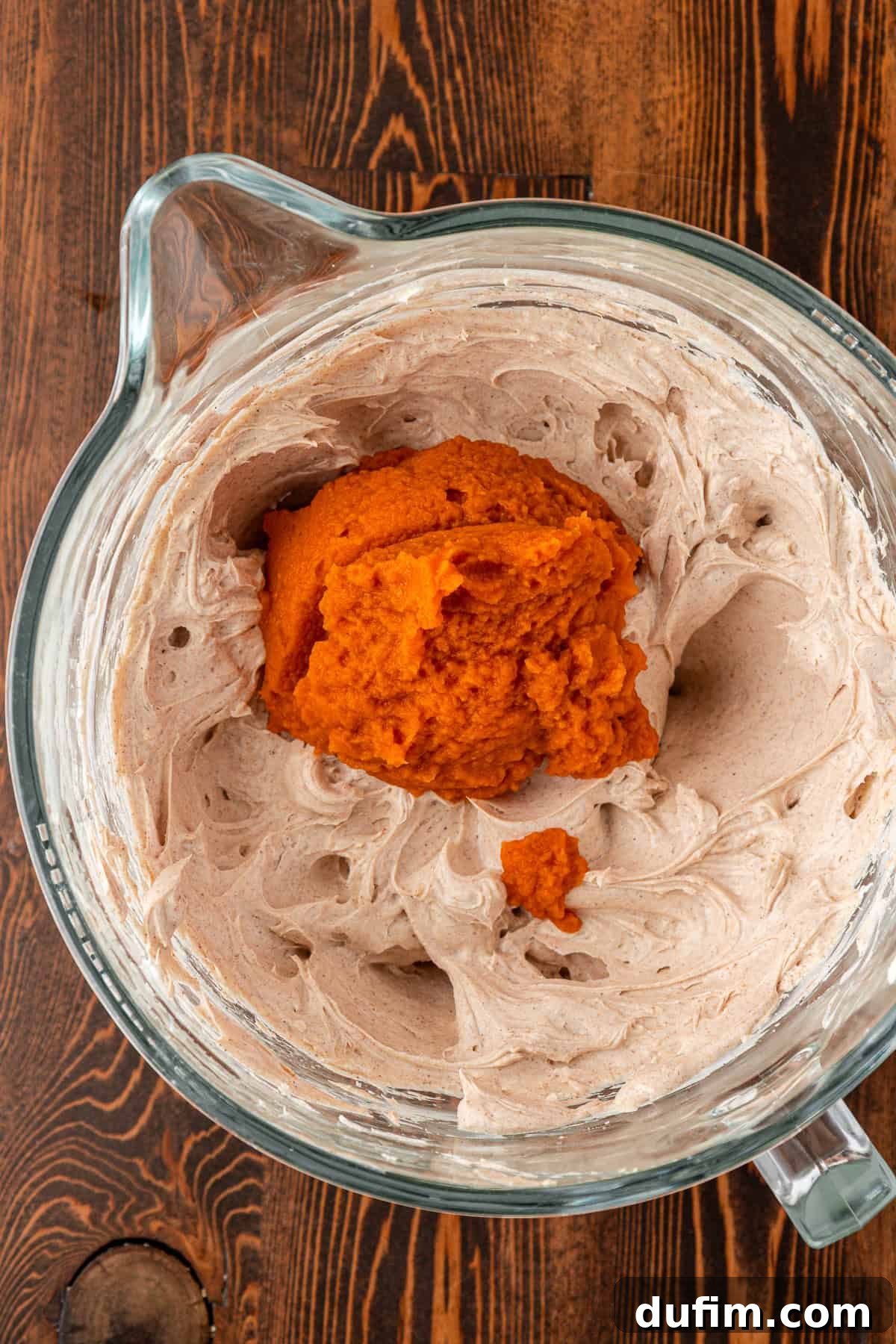 Spiced Pumpkin Cheesecake Squares 14 A glass mixing bowl on a wooden surface contains creamy whipped mixture with a mound of bright orange pumpkin puree on top, ready to be blended into the cheesecake batter.
