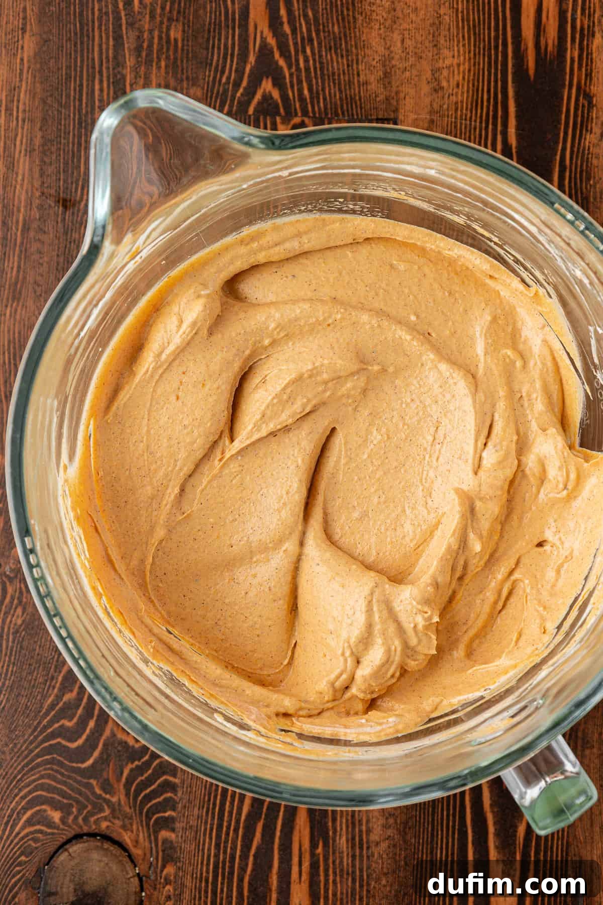Spiced Pumpkin Cheesecake Squares 15 A glass mixing bowl filled with creamy, light brown batter sits on a wooden surface. The batter is smoothly blended with swirls visible on top, indicating perfect incorporation of pumpkin puree.