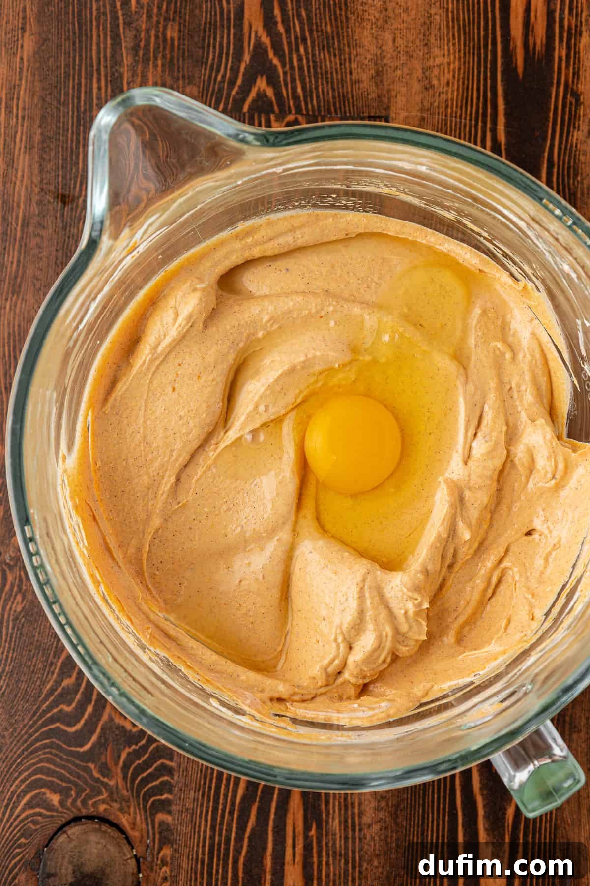 Spiced Pumpkin Cheesecake Squares 16 A glass mixing bowl with creamy light brown batter and a raw egg on top, set on a wooden surface, ready to be gently mixed.