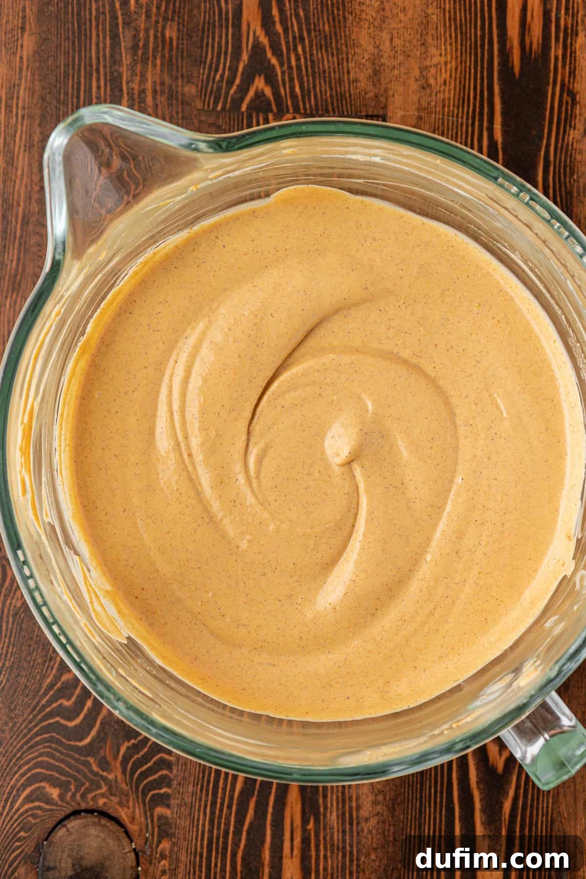 Spiced Pumpkin Cheesecake Squares 17 A glass mixing bowl filled with creamy, light brown pumpkin pie filling, swirled smoothly on top, sitting on a wooden surface. The eggs are fully incorporated, creating a perfect cheesecake batter.