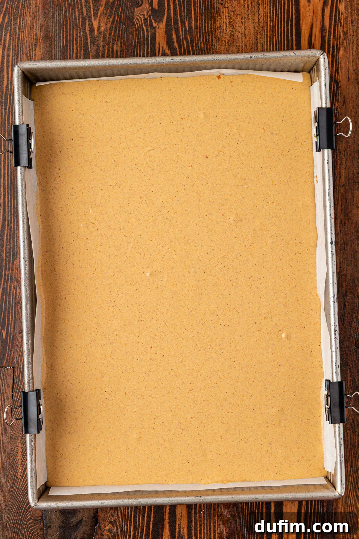 Spiced Pumpkin Cheesecake Squares 18 A parchment-lined square baking pan filled with a smooth, light brown batter, secured at the edges with binder clips, sits on a dark wooden surface. The pumpkin cheesecake mixture is ready for baking.