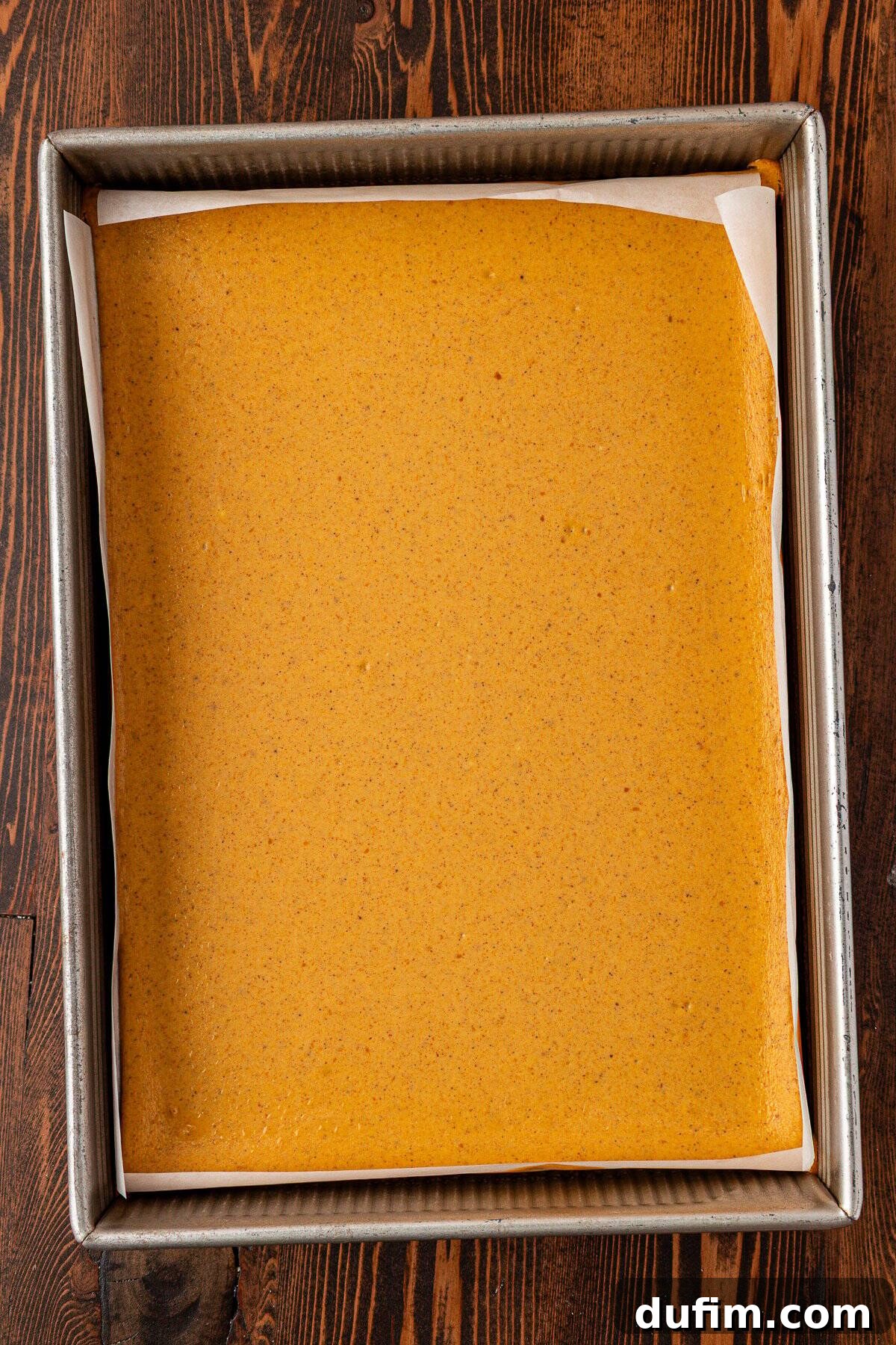 Spiced Pumpkin Cheesecake Squares 19 A rectangular baking pan lined with parchment paper, filled with a smooth, light brown batter, sits on a dark wooden surface. The surface of the cheesecake batter is even and perfectly prepared for baking.