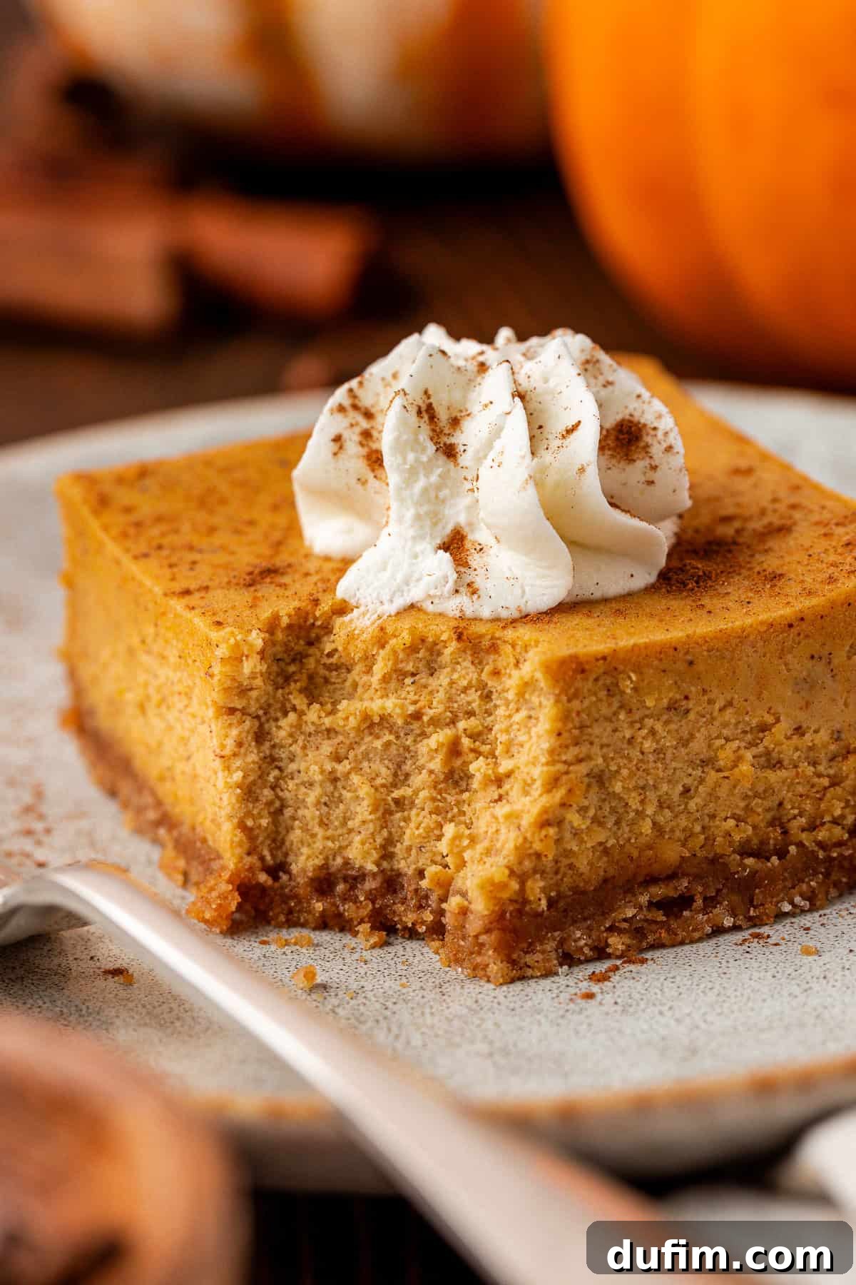 Spiced Pumpkin Cheesecake Squares 20 A square slice of pumpkin cheesecake with a graham cracker crust, topped with a swirl of whipped cream and sprinkled with cinnamon, sits on a plate with a fork. This perfectly baked pumpkin cheesecake bar is ready to be enjoyed.