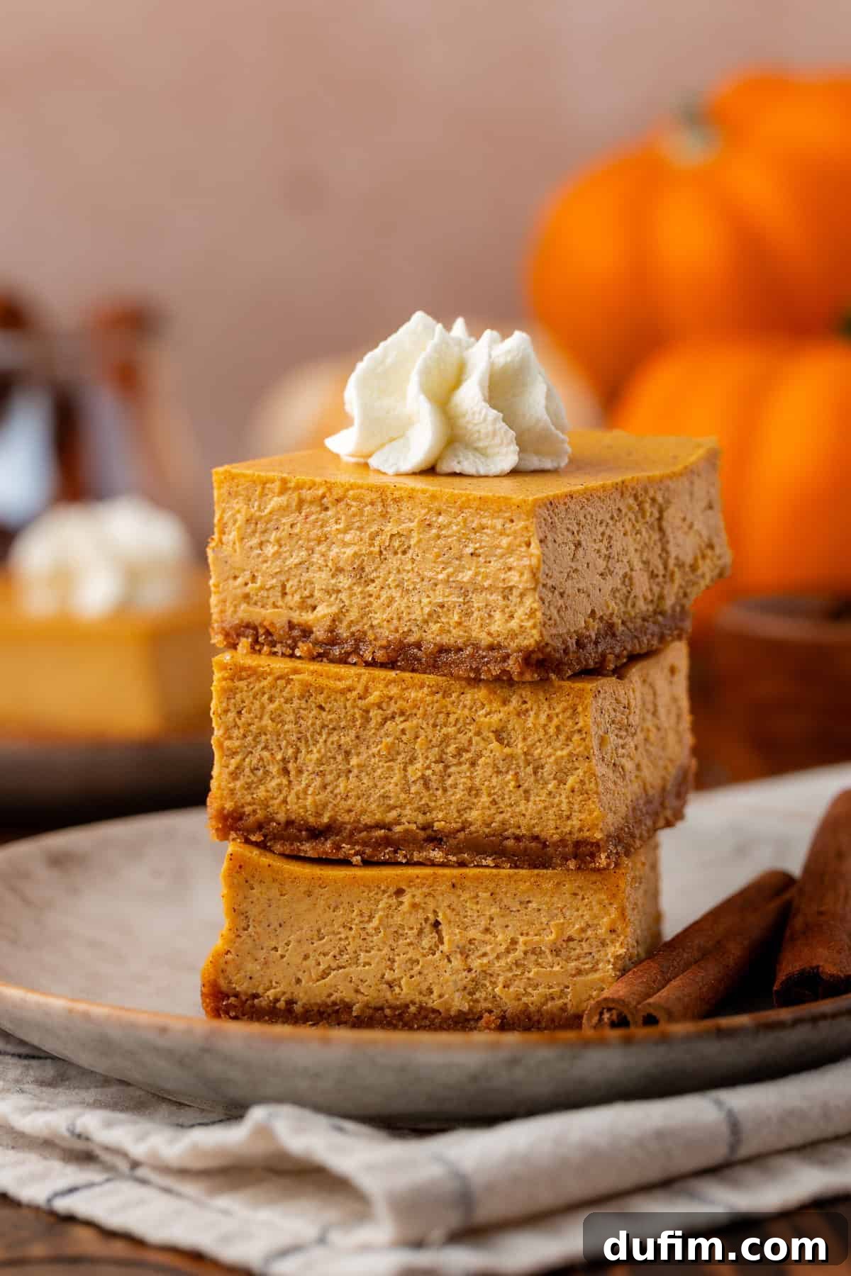 Spiced Pumpkin Cheesecake Squares 3 Three pumpkin cheesecake bars are stacked on a plate, topped with a swirl of whipped cream. Two cinnamon sticks lie beside the stack, and pumpkins are blurred in the background. Enjoy these creamy, spiced pumpkin cheesecake bars with a delightful graham cracker crust, a fuss-free dessert for any occasion.