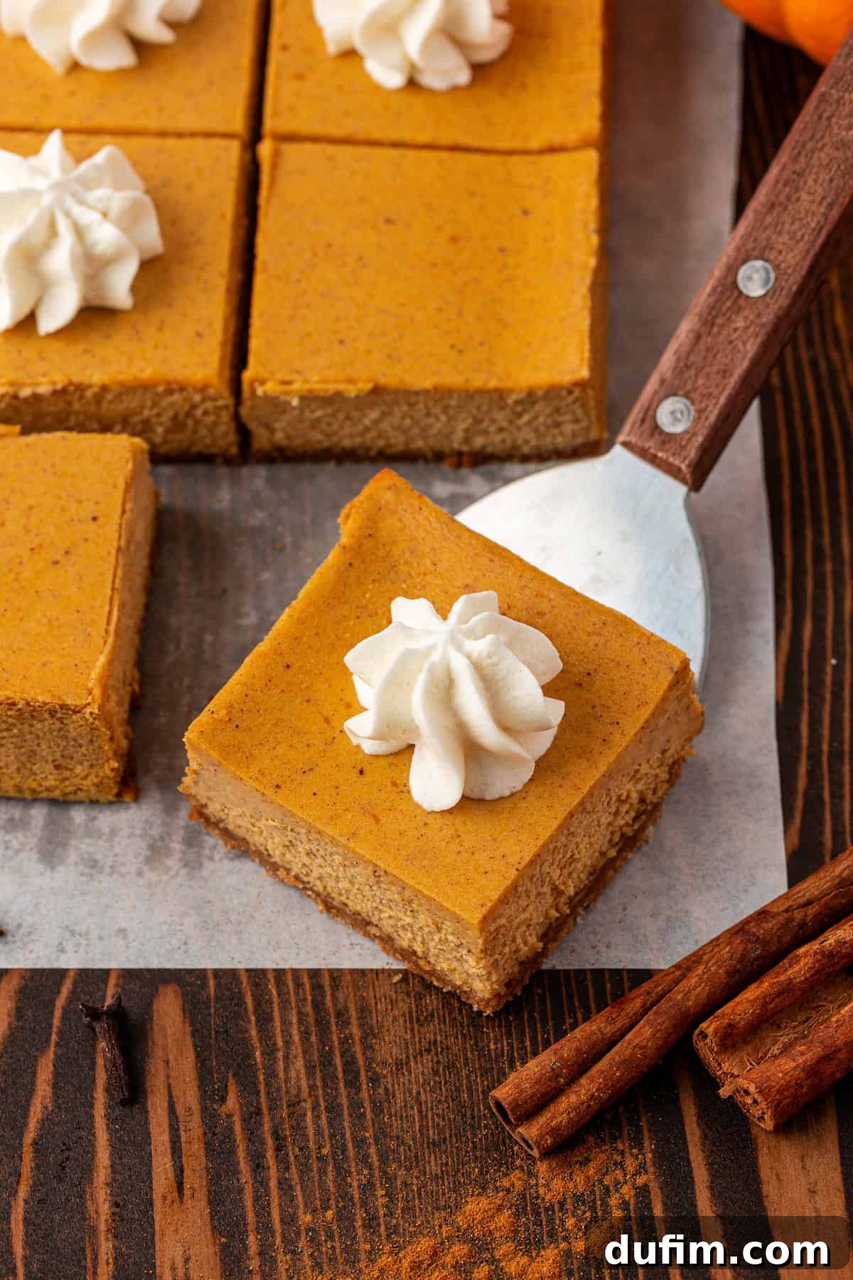 Spiced Pumpkin Cheesecake Squares 22 A close-up of pumpkin bars topped with whipped cream, arranged on parchment paper. One bar is being lifted with a spatula. Cinnamon sticks and a dusting of cinnamon are on the wooden table, highlighting the perfect serving of these creamy pumpkin cheesecake bars.