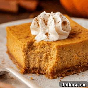 A square slice of pumpkin cheesecake with a graham cracker crust, topped with whipped cream and a sprinkle of cinnamon, sits on a plate with a bite taken out.