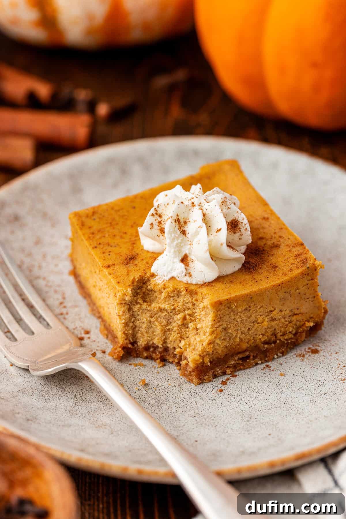 Spiced Pumpkin Cheesecake Squares 4 A square slice of pumpkin cheesecake with a crumbly crust, topped with a swirl of whipped cream and a sprinkle of cinnamon, sits on a plate next to a fork. Pumpkins and cinnamon sticks are in the background. This perfect slice of pumpkin cheesecake bar offers creamy, spiced flavor without the fuss.