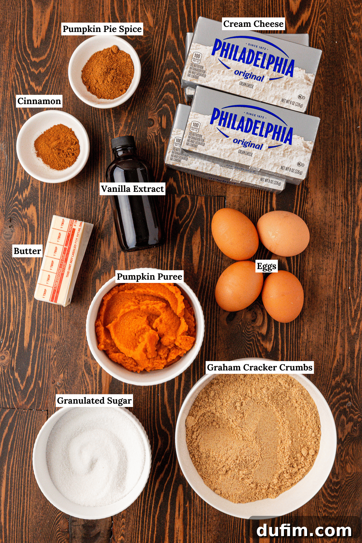Spiced Pumpkin Cheesecake Squares 5 Ingredients for pumpkin cheesecake on a wooden table: cream cheese, pumpkin puree, granulated sugar, graham cracker crumbs, eggs, butter, vanilla extract, pumpkin pie spice, and cinnamon, each labeled. These simple ingredients combine to create the best easy pumpkin cheesecake bars.