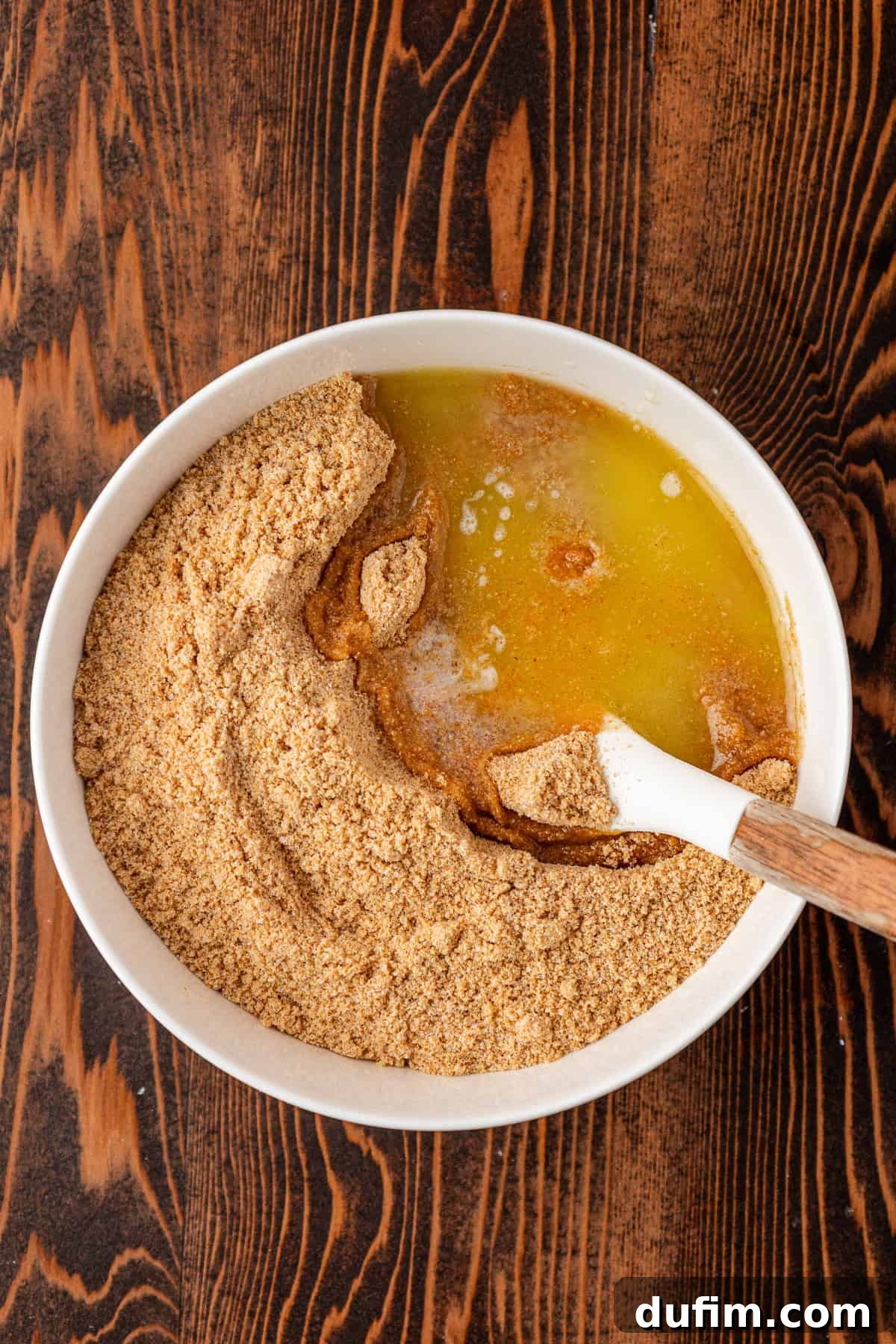 Spiced Pumpkin Cheesecake Squares 6 A white bowl filled with graham cracker crumbs and melted butter sits on a wooden table, with a spatula partially mixing the ingredients, ready for a delicious crust.