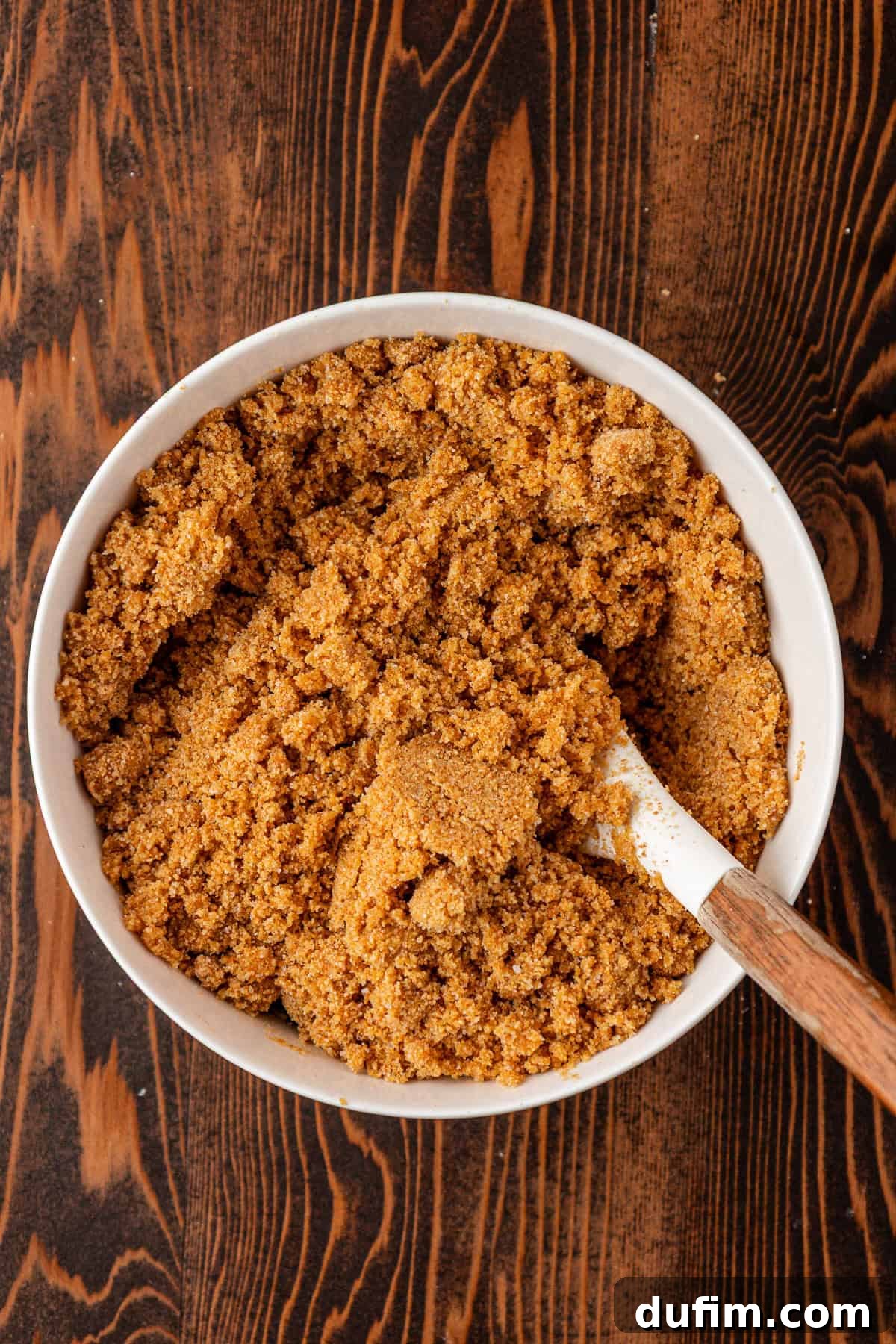Spiced Pumpkin Cheesecake Squares 7 A white bowl filled with crumbly, golden brown graham cracker crust mixture sits on a dark wooden surface, with a wooden-handled spatula resting inside the bowl. The perfect texture for a cheesecake base.