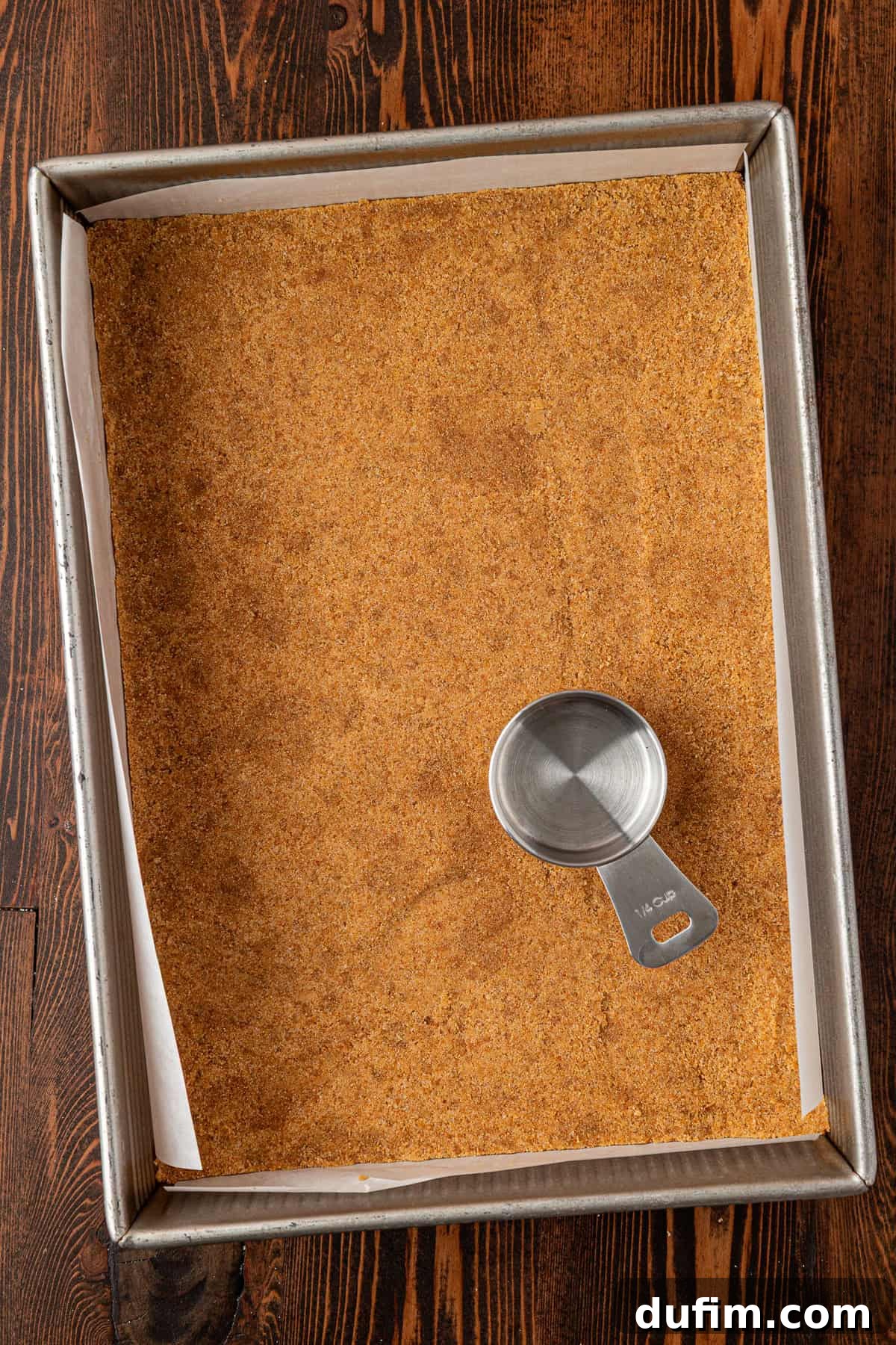 Spiced Pumpkin Cheesecake Squares 8 A baking pan lined with parchment paper containing a pressed graham cracker crust, with a metal measuring cup resting on the surface. The pan is on a dark wooden table, ensuring an even and sturdy crust base.