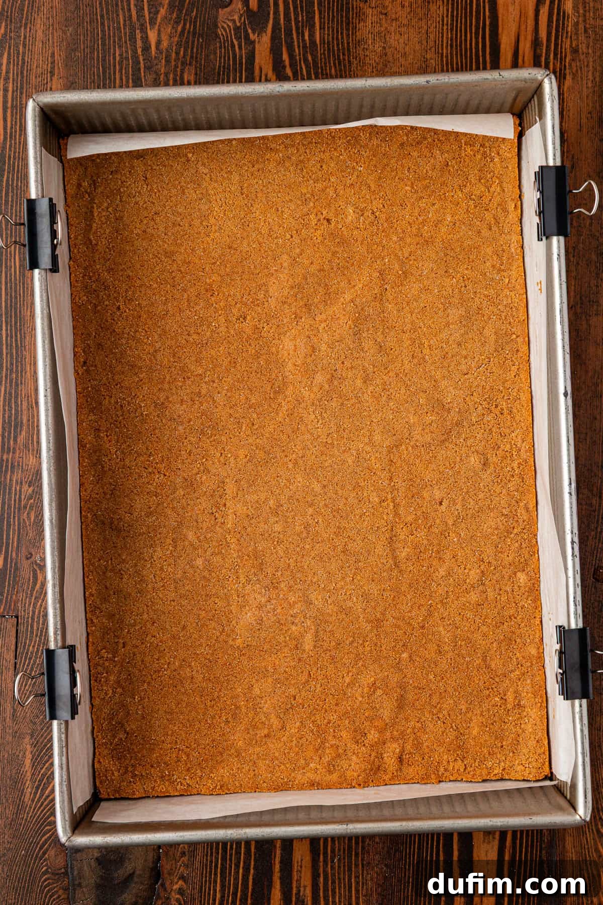 Spiced Pumpkin Cheesecake Squares 9 A square baking pan lined with parchment paper and binder clips holds a flat, golden-brown graham cracker crust on a wooden table. The crust is evenly pressed and ready for the cheesecake filling.