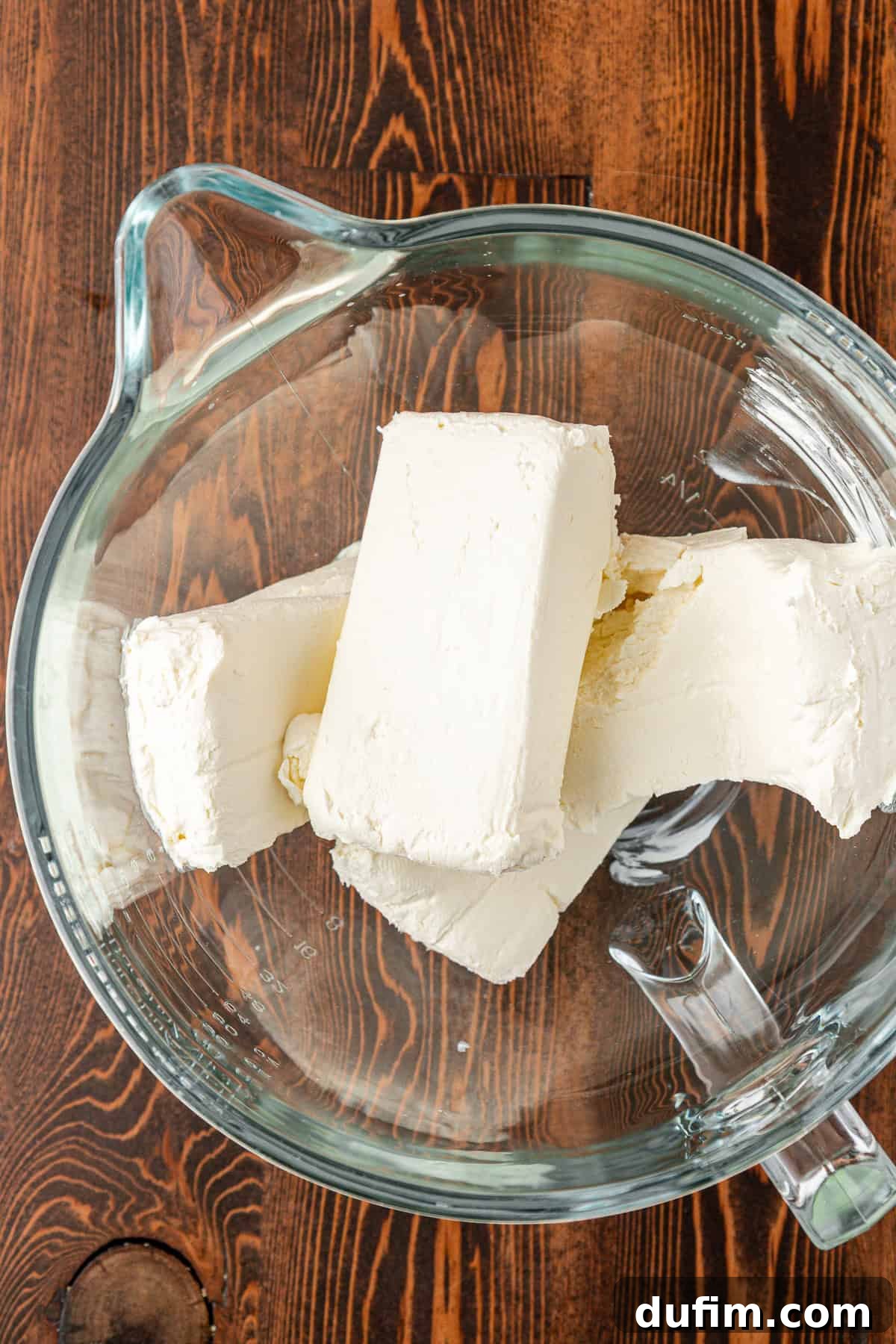 Spiced Pumpkin Cheesecake Squares 10 Three blocks of cream cheese in a clear glass mixing bowl, placed on a wooden surface. Softened cream cheese is essential for a smooth cheesecake filling.